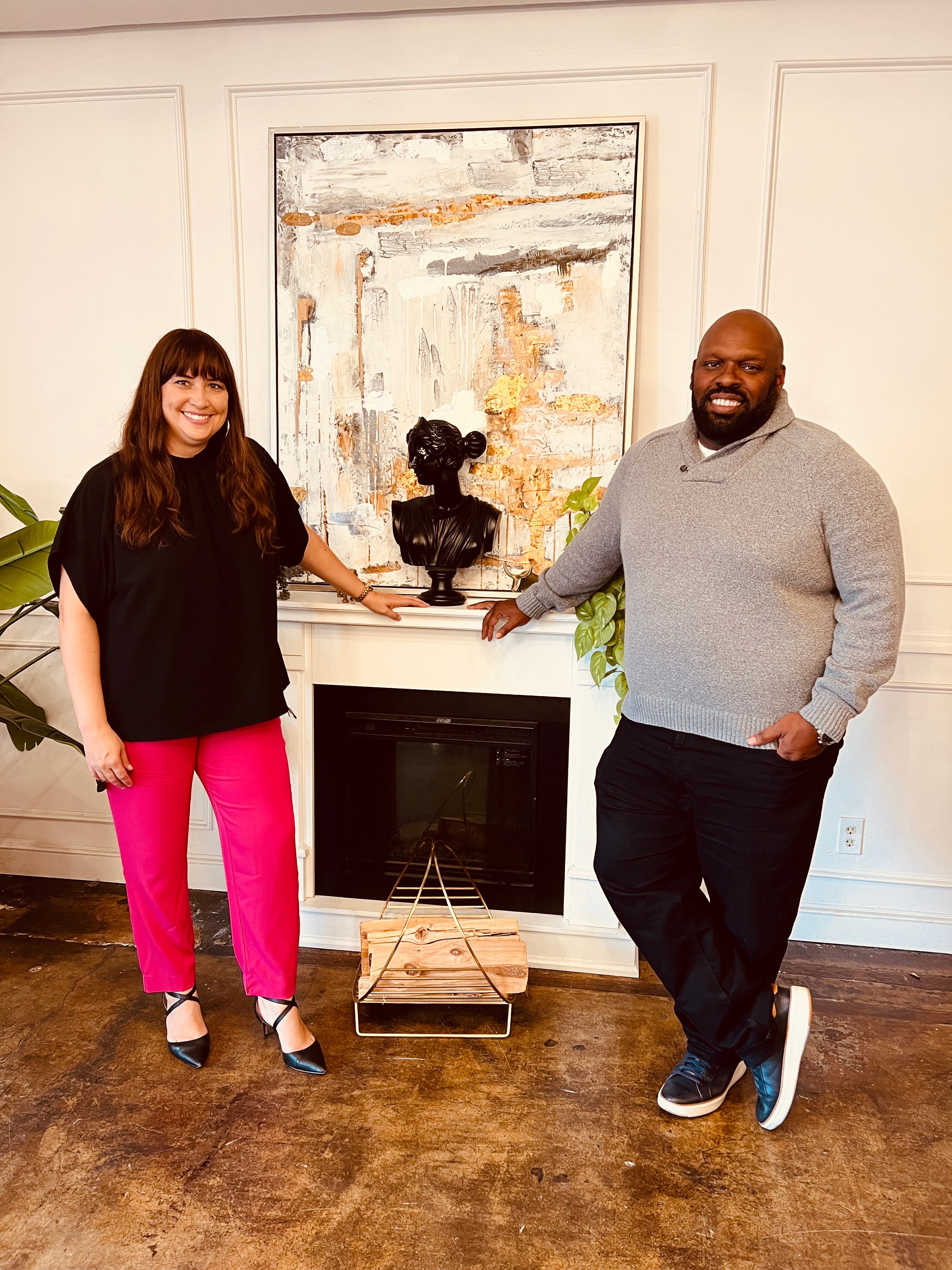 Two people, the founders, stand in front of fire[lace smiling. The woman is on the left. She is wearing a black top and bright pink long pants with black heels. The man on the right is wearing a grey sweater, black pants, and sneakers. 